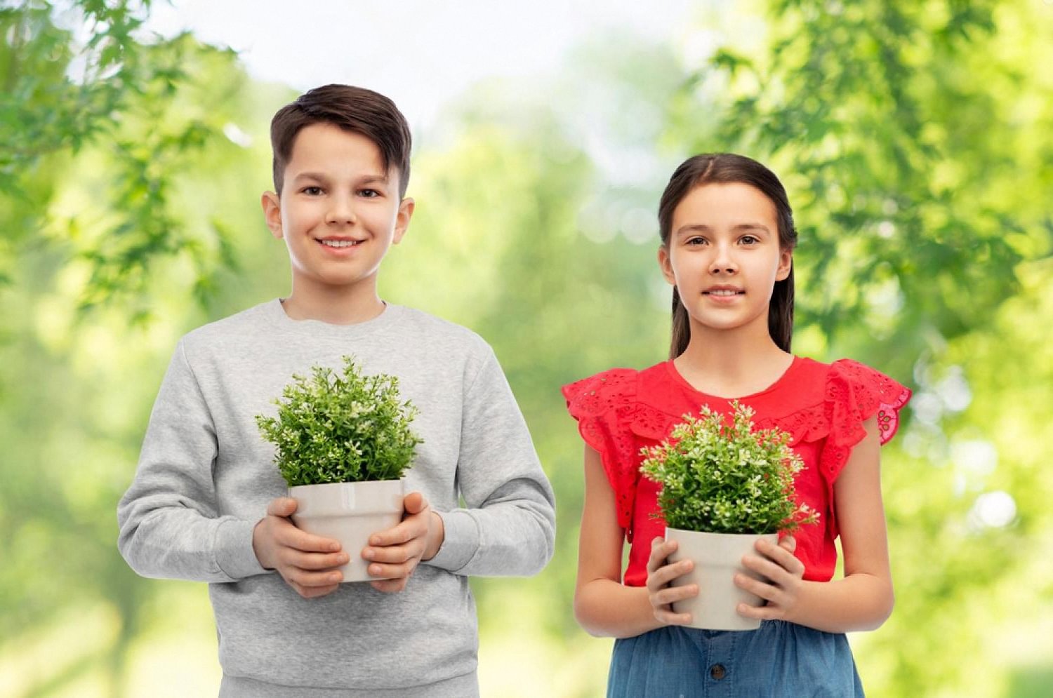 Boy and girl in school clothes holding potted plants in front of green foliage